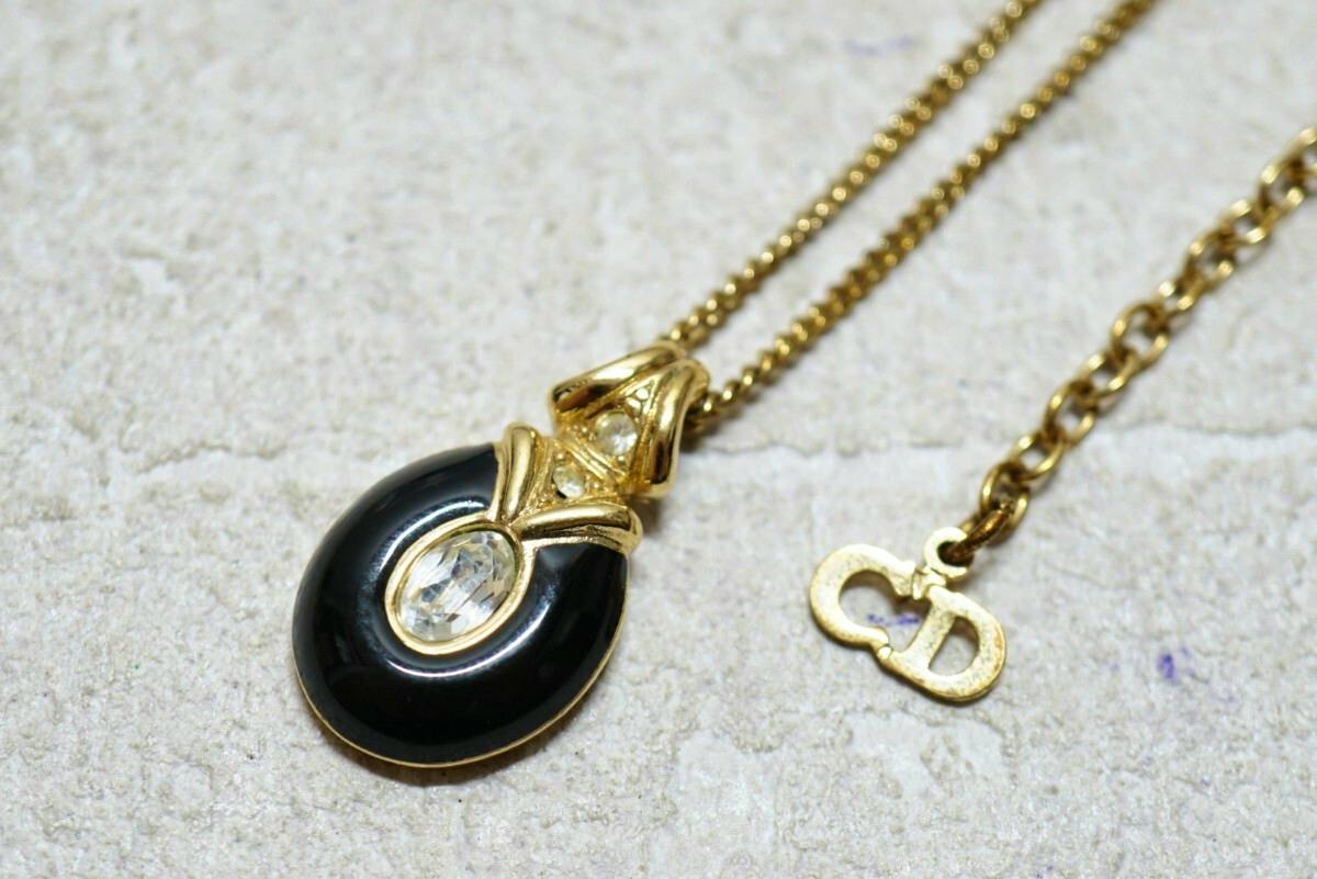 Close-up of the Christian Dior gold-color necklace showing a black teardrop pendant with a clear stone and a small 'CD' logo charm on a textured surface.