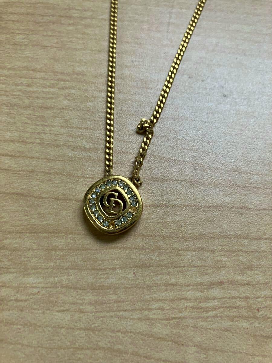 Gold-tone Christian Dior necklace with a rhinestone-studded round pendant bearing a CD logo on a wood background.