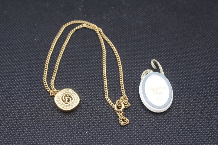 Gold-tone Christian Dior necklace with a round pendant bearing a CD logo and chain displayed with a white oval tag on a black textured background.