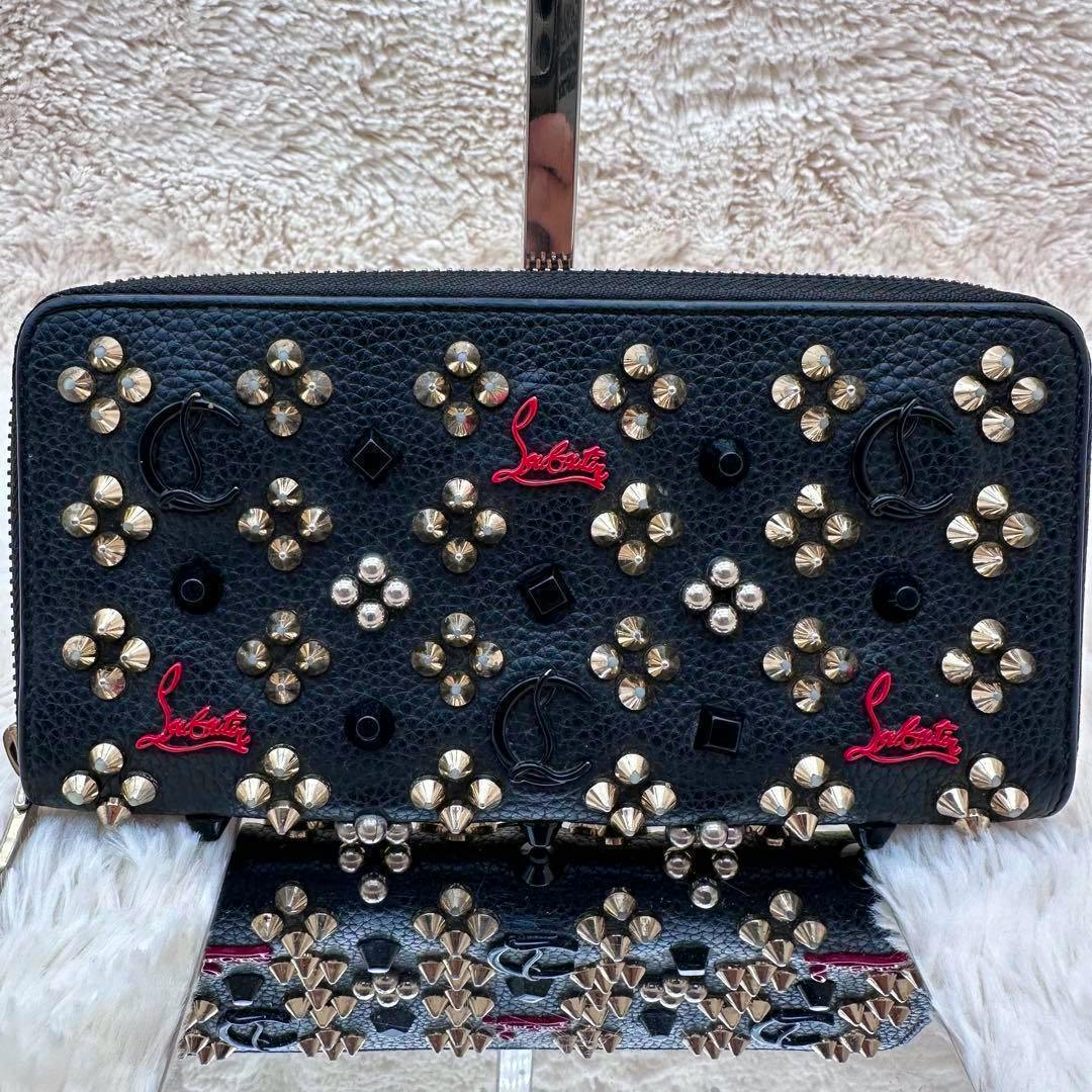 Front view of a black pebbled leather zip-around long wallet decorated with metal studs and red Louboutin logos.