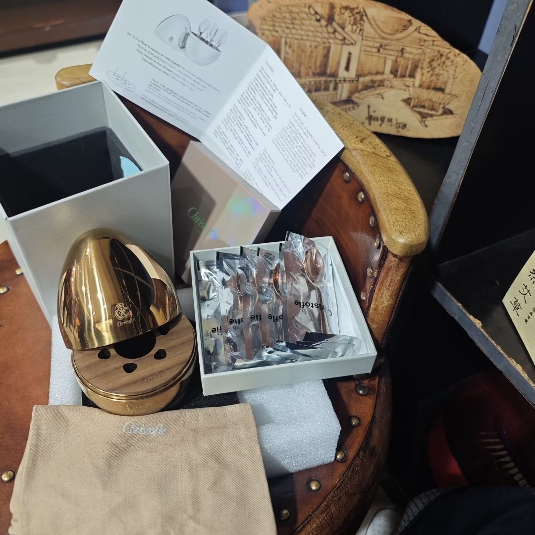 Open box showing the Christofle Mood rose gold coffee spoon 6-piece set and accessories on a leather chair.