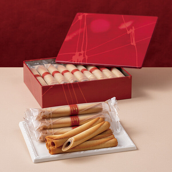 Opened red gift box of シガール オゥ ショコラ showing individually wrapped cigar-shaped cookies and several unwrapped cookies on a white plate.