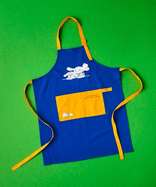 Blue apron with a yellow pocket and yellow straps featuring the アイシナモロール character print, laid flat on a green background.