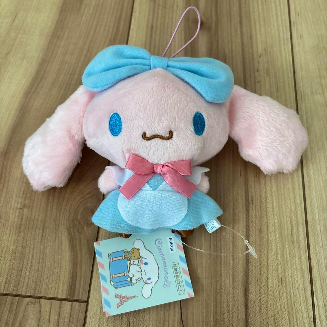 Front view of the Cinnamoroll mascot plush in pink wearing a blue dress and pink bow, showing its face, hanging loop, and attached tag on a wood floor.
