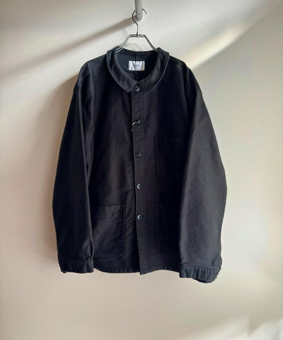 Black CIOTA moleskin French work jacket hanging on a hanger against a plain wall, front view showing buttons and pockets.