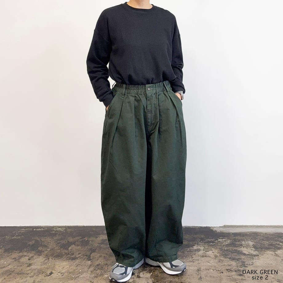 Front view of a model wearing dark green circus pants with pleated wide legs and an elasticated waistband.