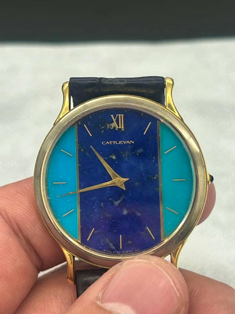 Front view of the CATTLEYAN watch showing a blue and turquoise dial with stick indices and gold-tone hands held between fingers.