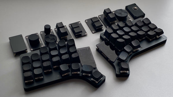 Split ergonomic PC keyboard (Claw44 v3) with black keycaps, rotary knobs, and module plates on a gray surface.
