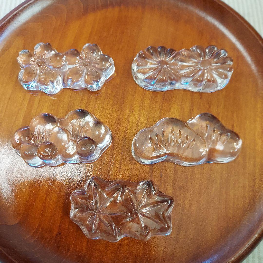 Five transparent glass chopstick rests shaped like flowers and leaves on a wooden plate.
