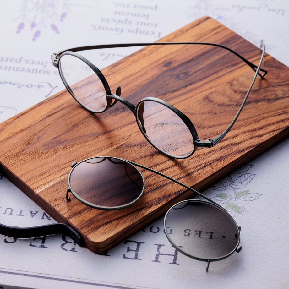 Round metal eyeglasses with two detachable round clip-on sunglass lenses displayed on a wooden board.