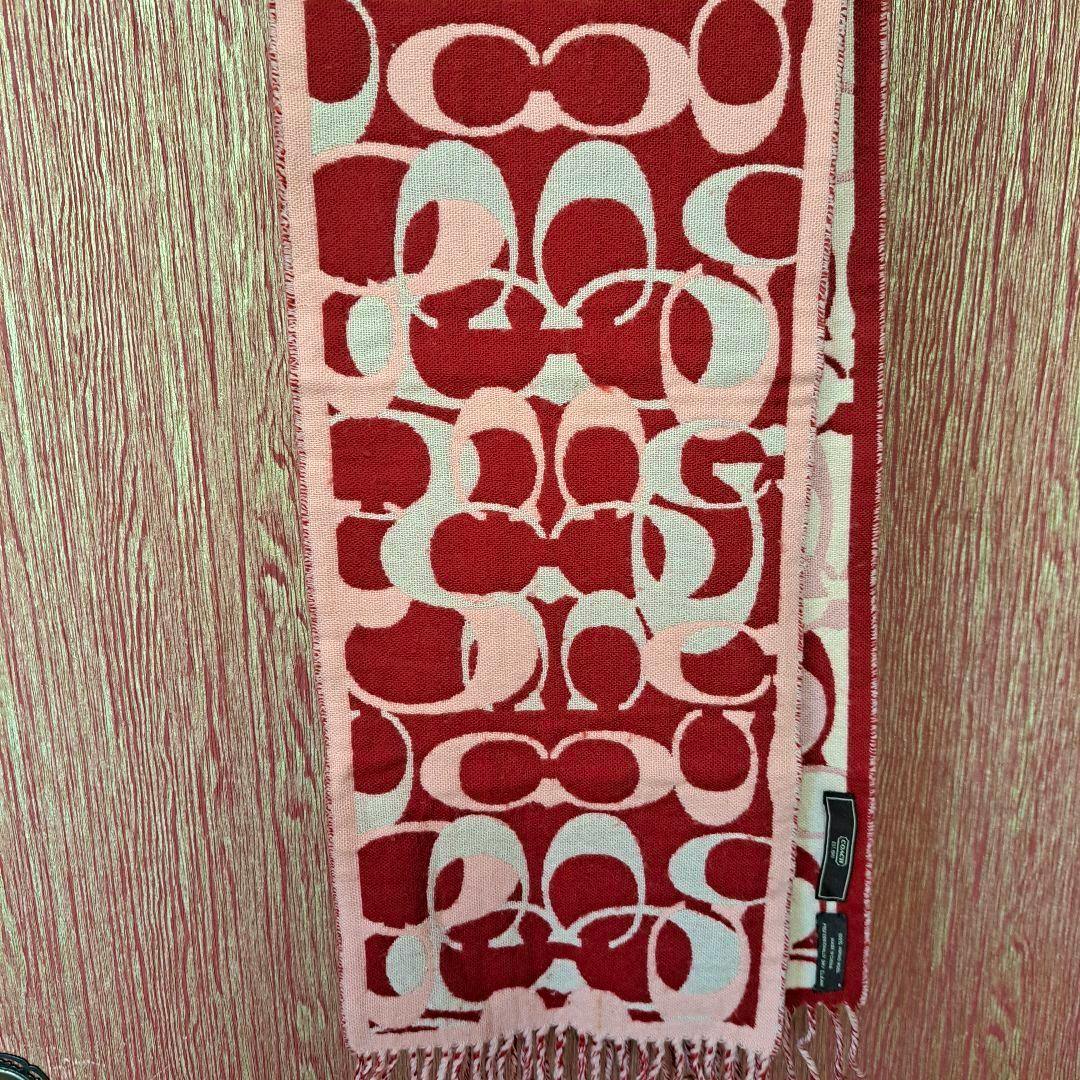 A red and pink COACH wool scarf with signature 'C' pattern hung vertically against a textured background.