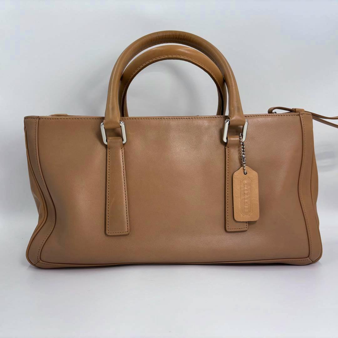 Front view of a tan leather Coach tote handbag with two top handles and a leather hang tag against a white background.