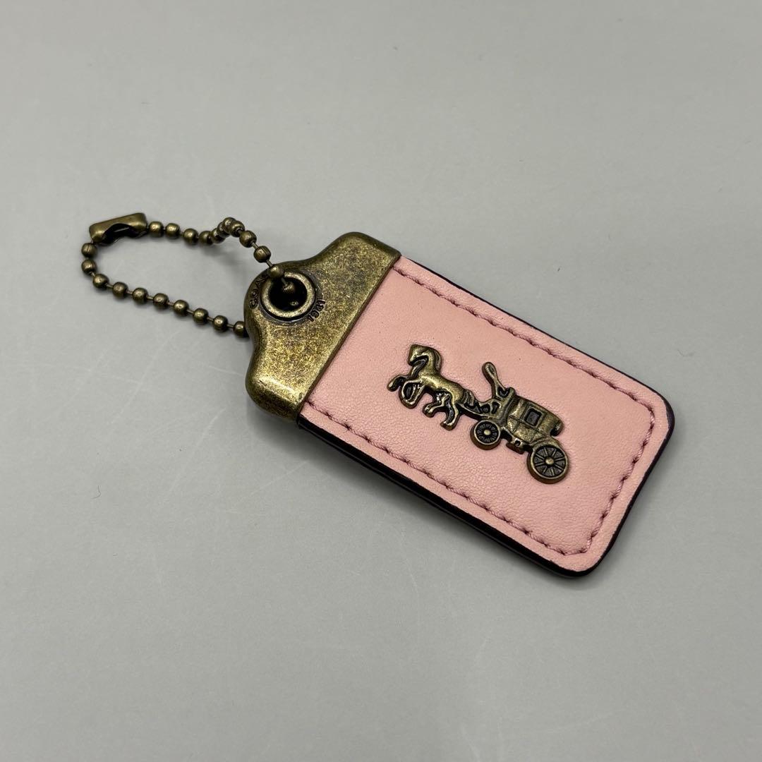 Pink cow-leather COACH hangtag keychain with an antiqued metal top and horse-and-carriage emblem on a gray background.