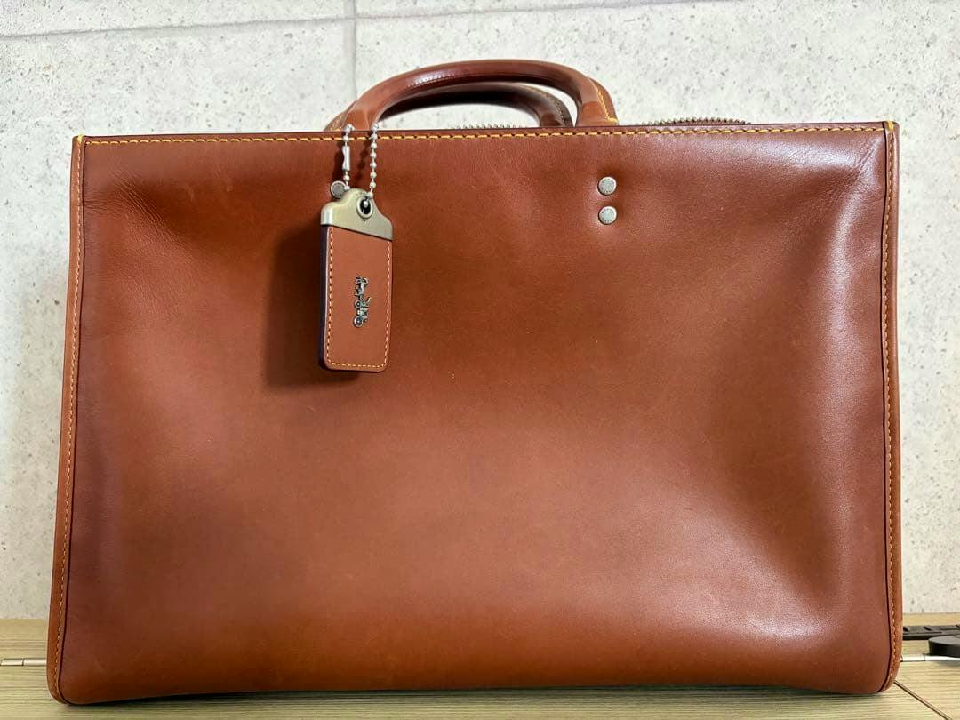 Front view of the COACH 1941 Rogue brief leather business bag showing the front panel, handles, and leather hang tag.
