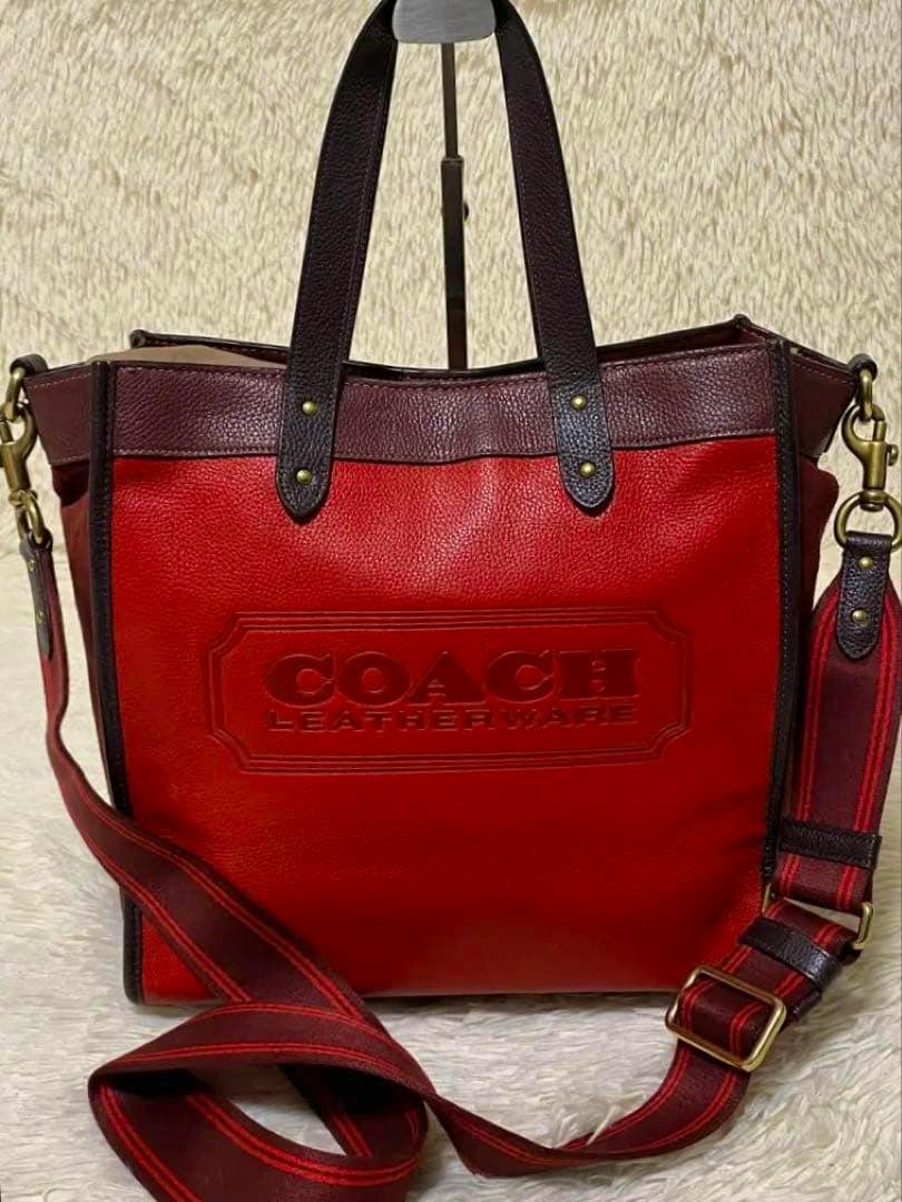 Front view of a red COACH 2-way pebble leather tote bag with dark brown handles and a detachable red-striped shoulder strap.