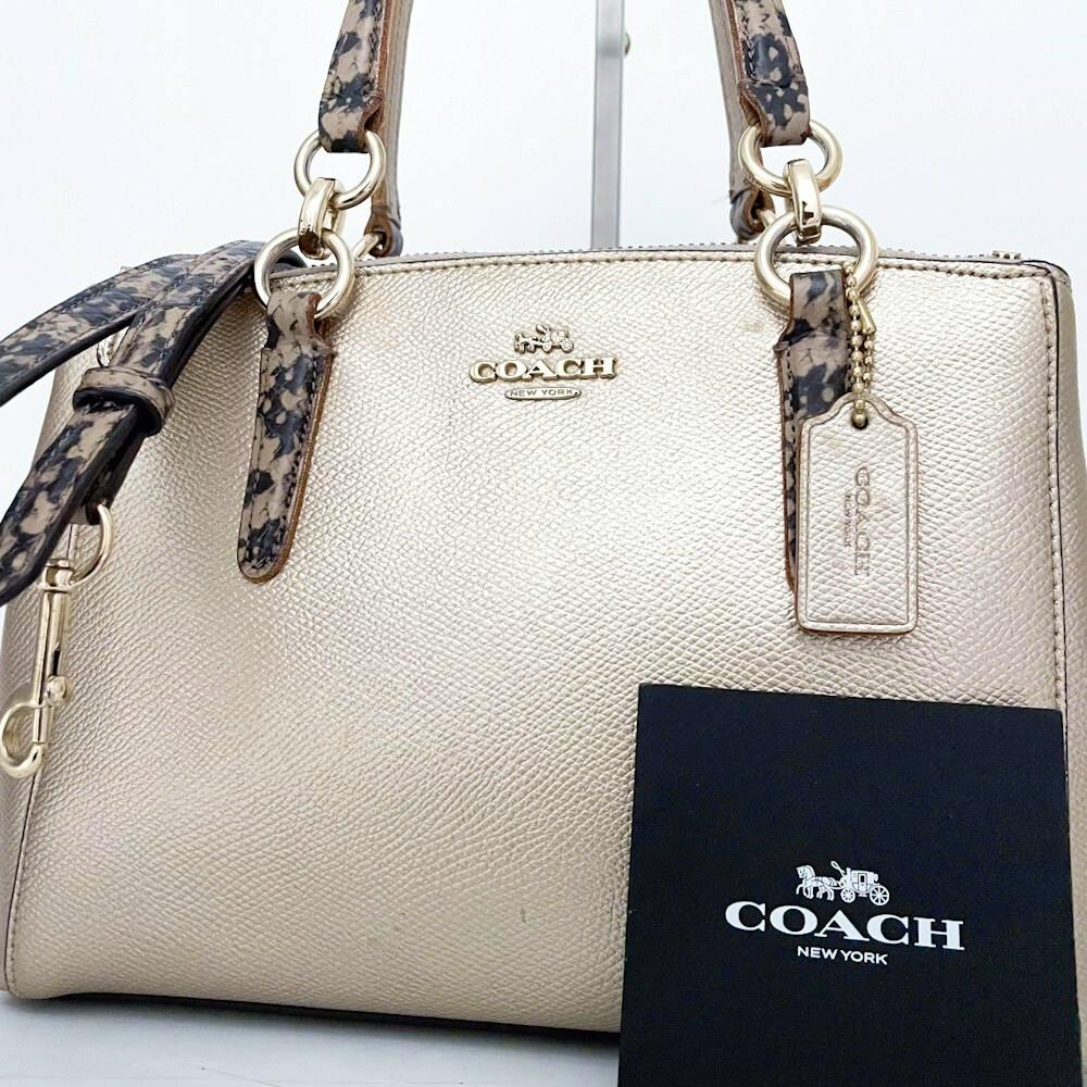 Front view of the COACH beige-gold tote bag showing patterned straps, a leather hang tag, and a COACH card.
