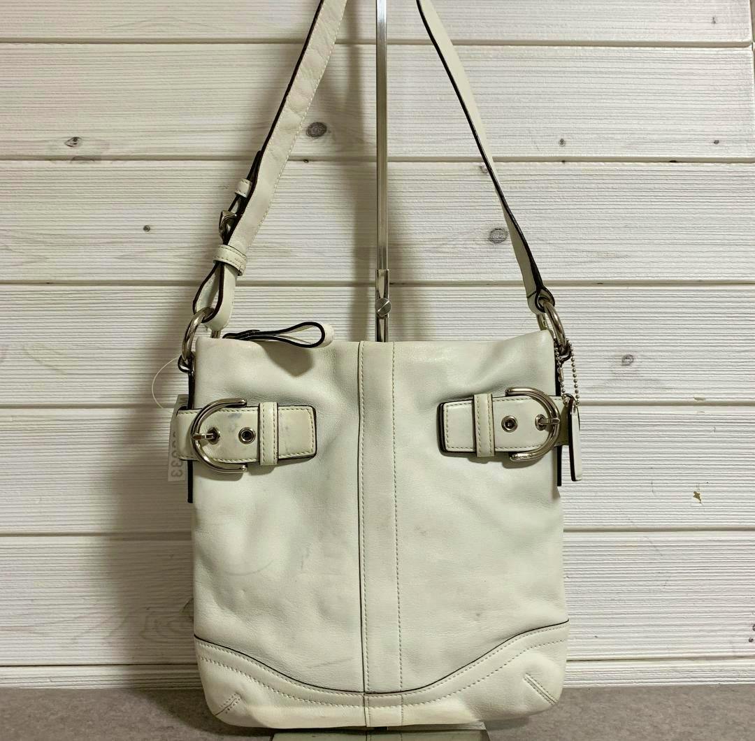 White Coach cow leather crossbody shoulder bag with buckle accents and a shoulder strap hanging against a wood-paneled wall.
