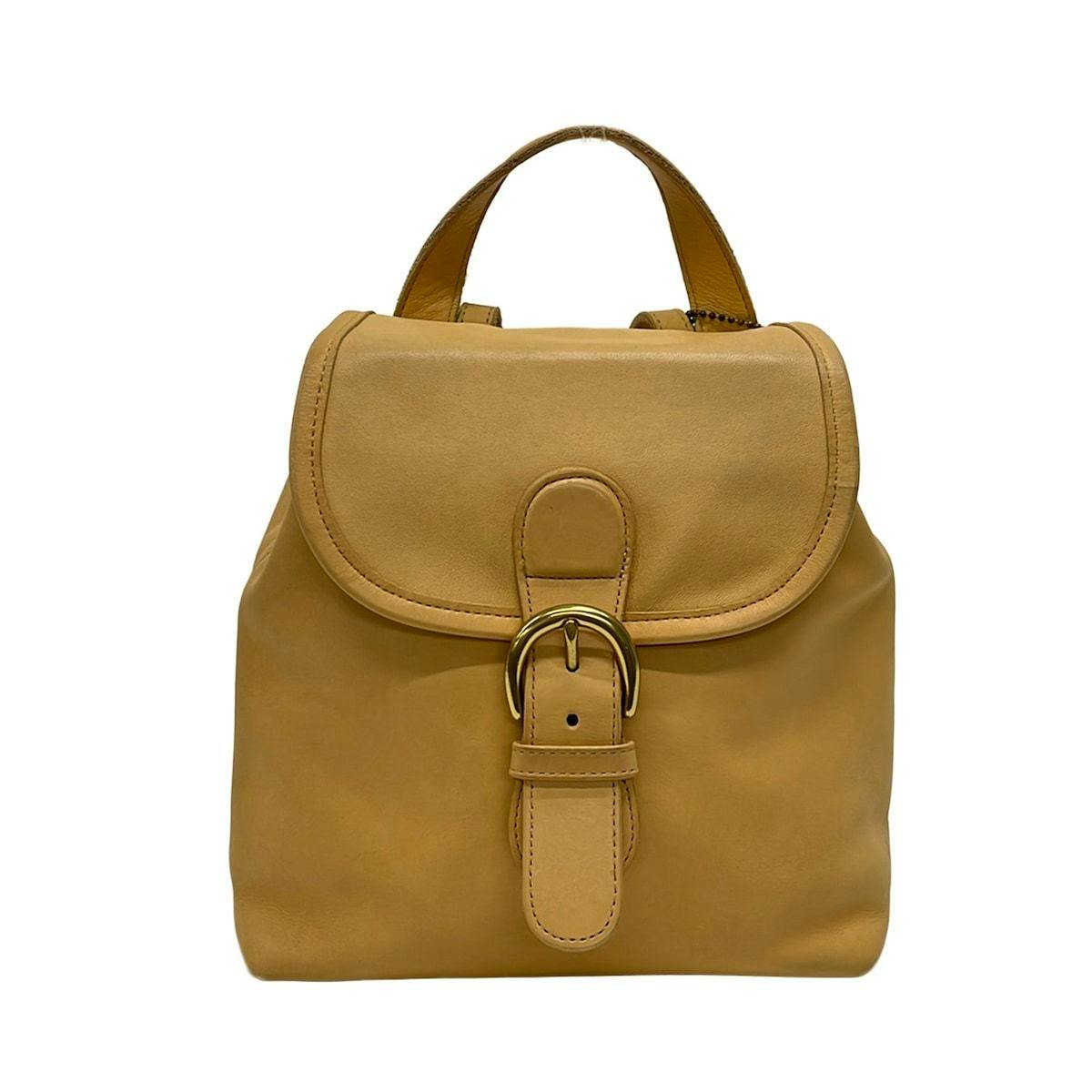 Front view of a beige COACH leather backpack showing a top handle and a buckle flap closure.