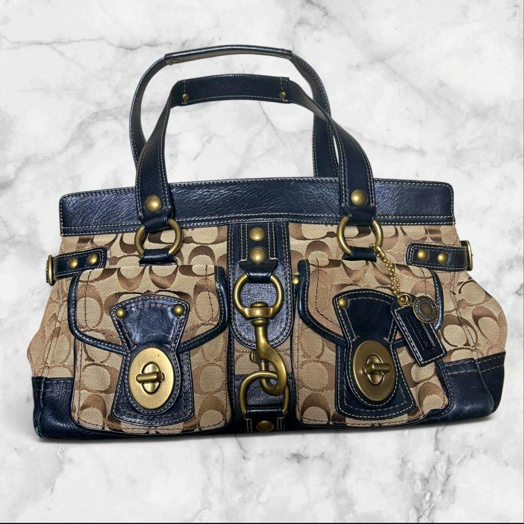 Front view of the COACH signature canvas bag with dark leather trim, front pockets, brass hardware, and a hanging Coach tag.