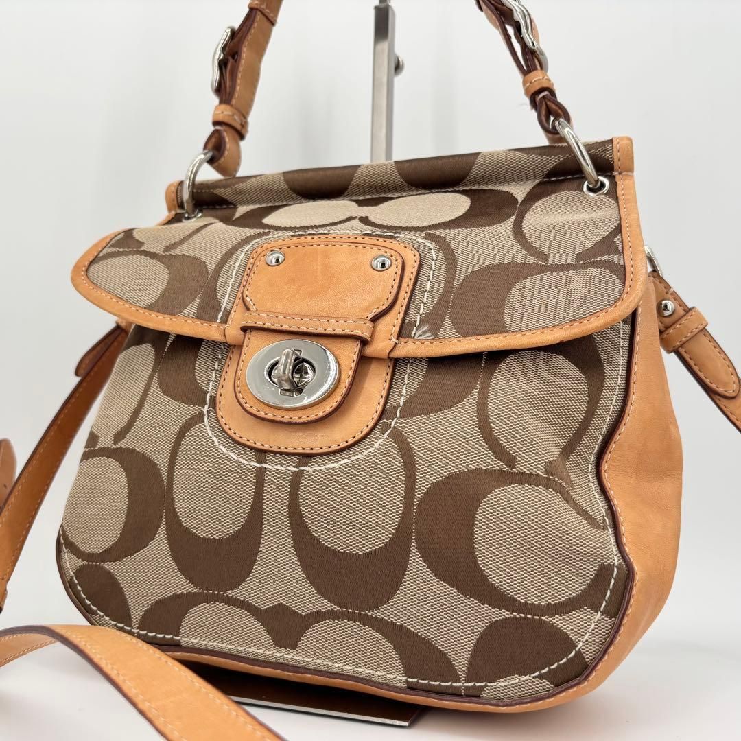 Front view of a Coach canvas signature shoulder/crossbody bag with a flap, turn-lock closure, and leather trim.