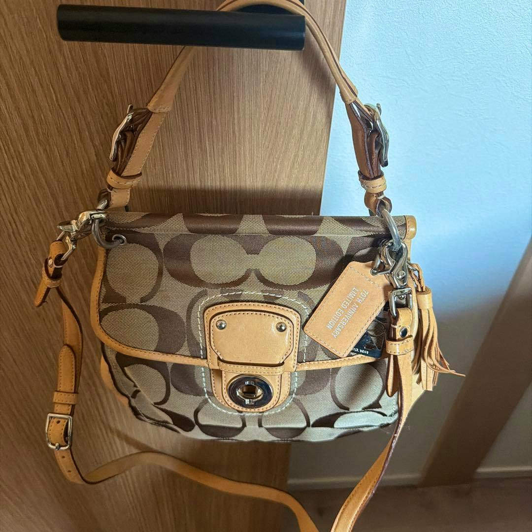 Front view of a COACH signature canvas shoulder bag with leather trim, a turn-lock closure, and a hanging leather tag.