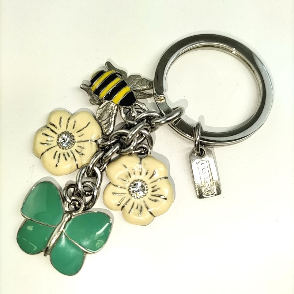 Photo of a COACH metal keychain showing a green butterfly charm, two cream flower charms, a yellow-and-black bee charm, and a small rectangular tag.