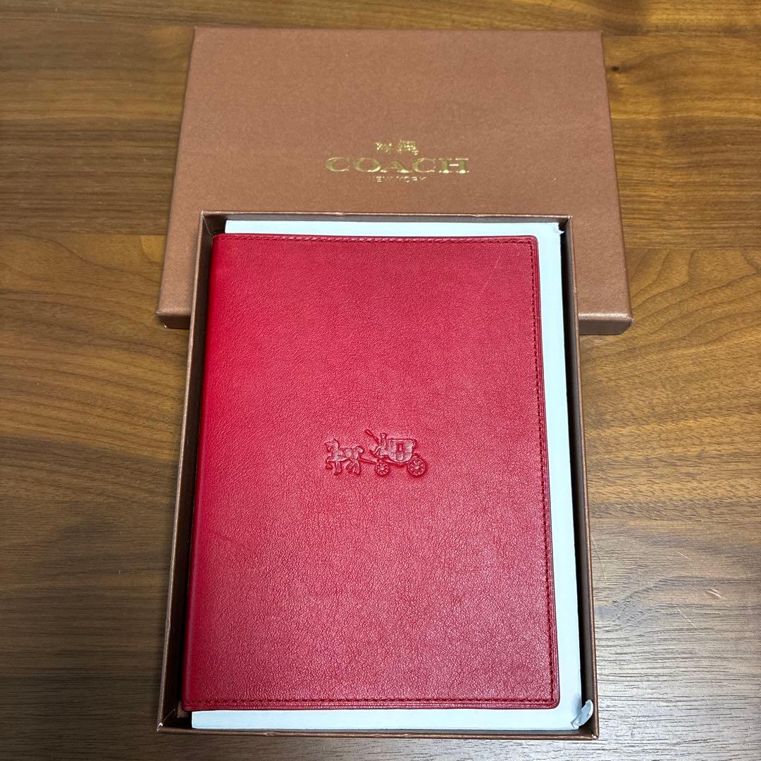 Red Coach leather A5 notebook cover with embossed logo shown inside its box on a wooden surface.