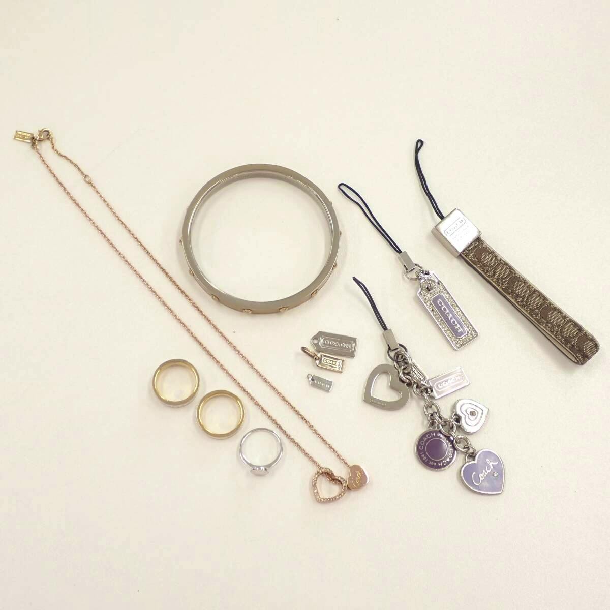 COACH accessories laid flat on a light background including a bangle, necklace, three rings, charm keychains, and a strap.