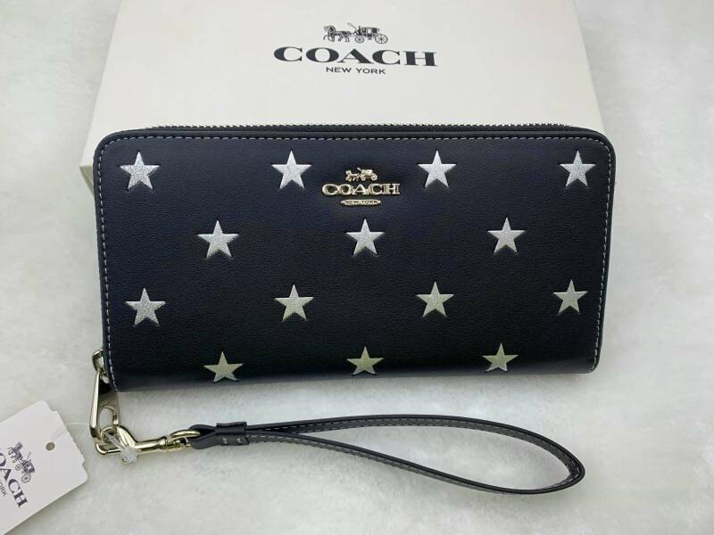 Black COACH zip-around long wallet with silver star pattern and a detachable wrist strap pictured on a white surface in front of a COACH box.