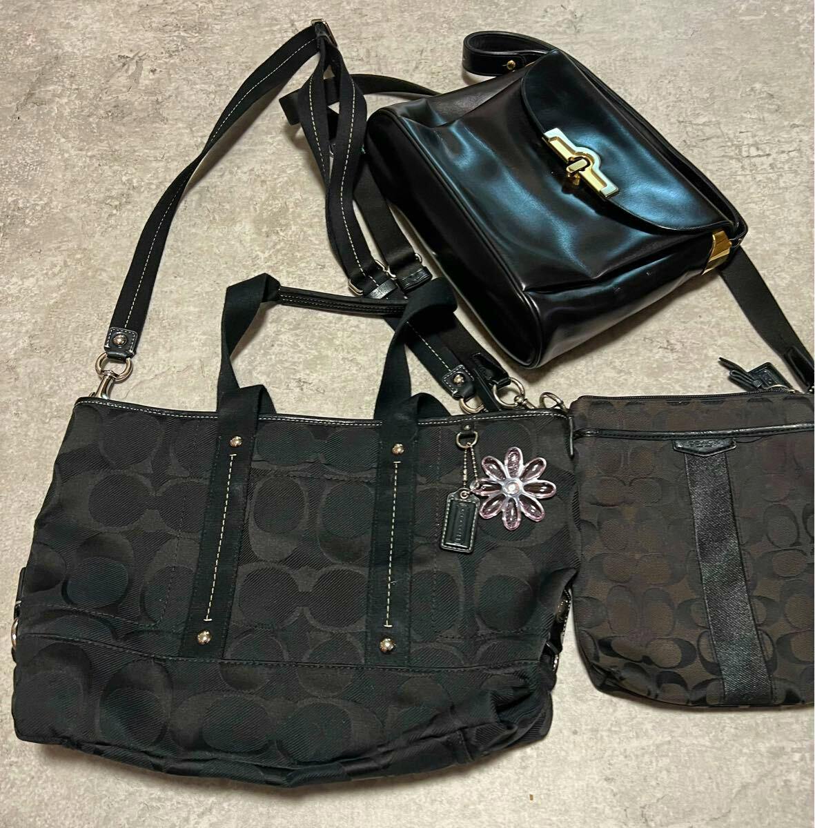 Three black bags (Coach and Bally) laid flat on a light floor, including two monogram canvas bags and one black shiny shoulder bag.