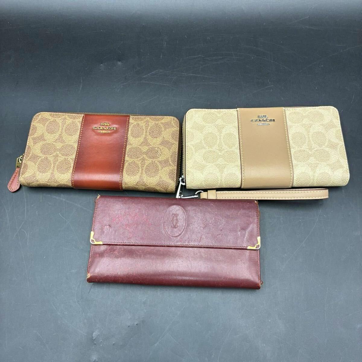 Three long wallets — two Coach canvas zip-around wallets and one Cartier burgundy leather wallet — arranged on a dark surface.