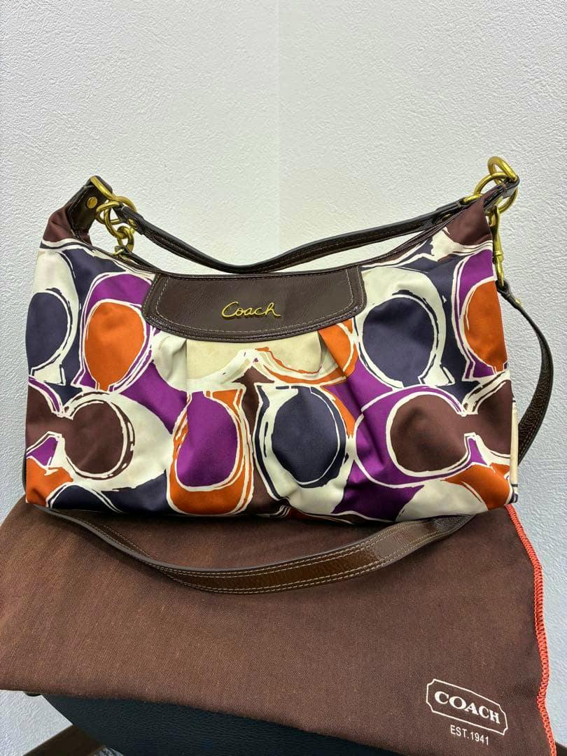 Front view of a Coach 2way shoulder bag with multicolor circular print, brown leather trim, and gold logo, shown on a Coach dust bag.