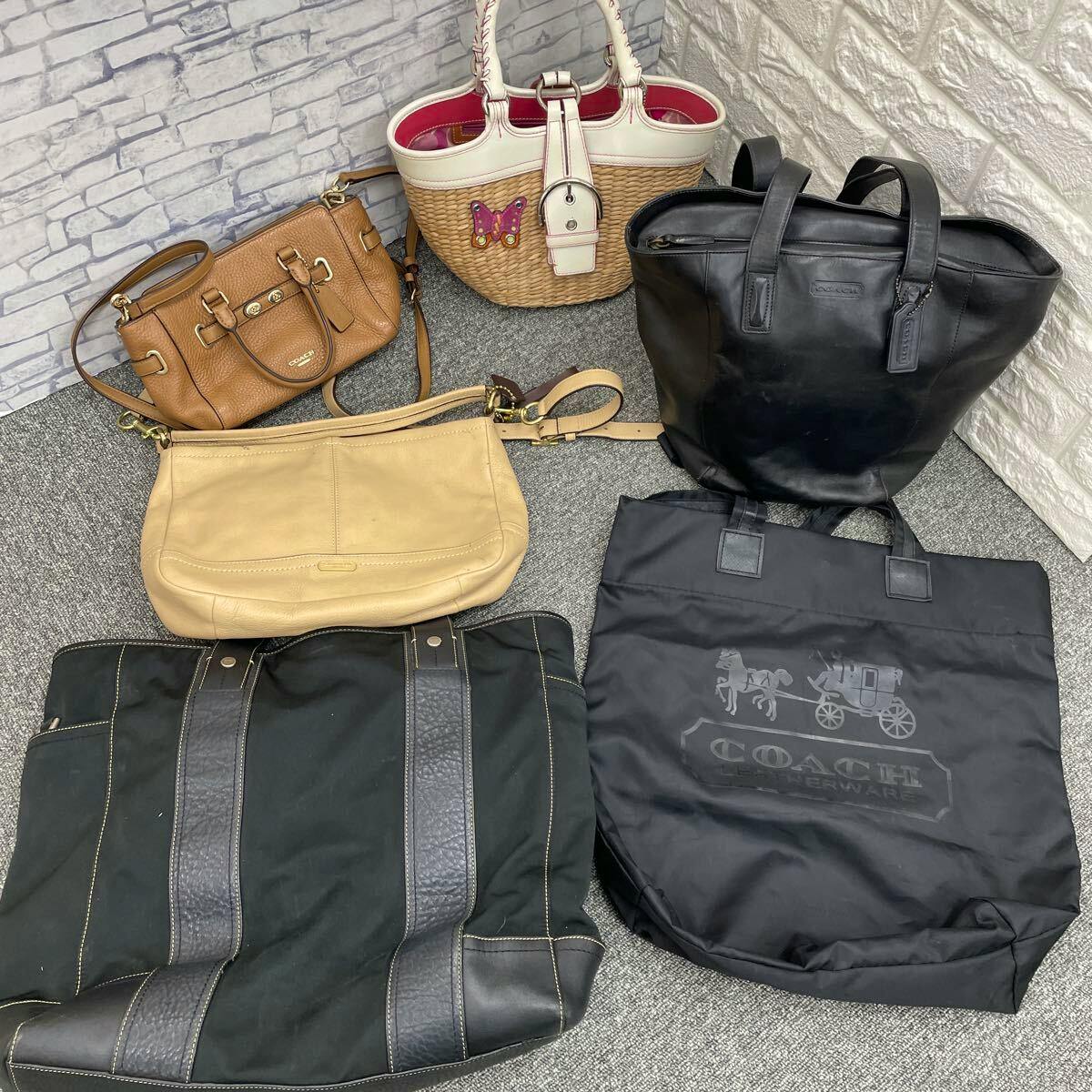 Six Coach bags of various styles and colors arranged on the floor, including a black tote, a straw basket bag with white trim, a brown handbag, a beige shoulder bag, and two black tote-style bags.