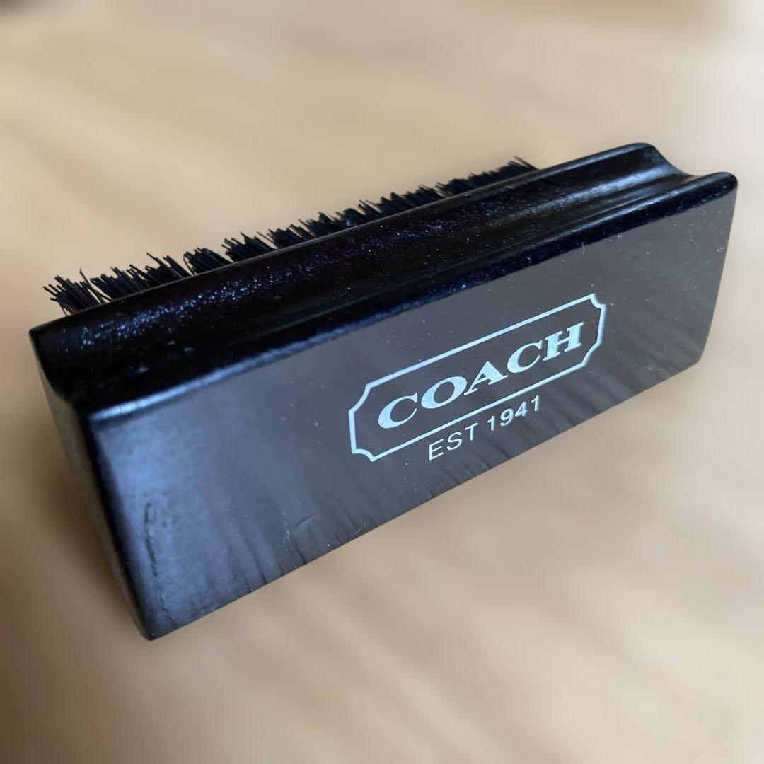 Close-up photo of a black COACH-branded bag care brush showing bristles and the COACH EST 1941 logo.