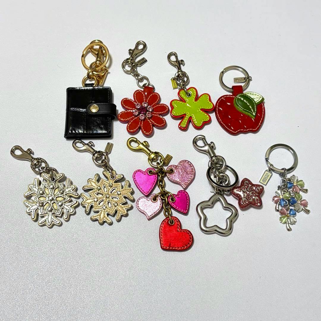 Nine COACH bag charms arranged on a white background, showing a black mini wallet, red flower, green clover, red apple, two snowflake pendants, a cluster of pink hearts, a metal star outline, a red star, and a pastel floral cluster.