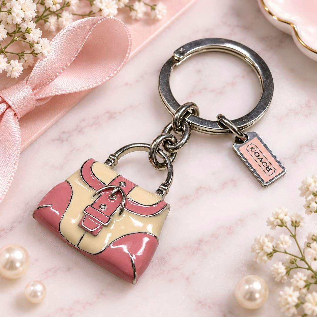 Pink bag-shaped COACH key ring with metal split ring and small COACH tag shown on a marble surface with decorative ribbon and pearls.