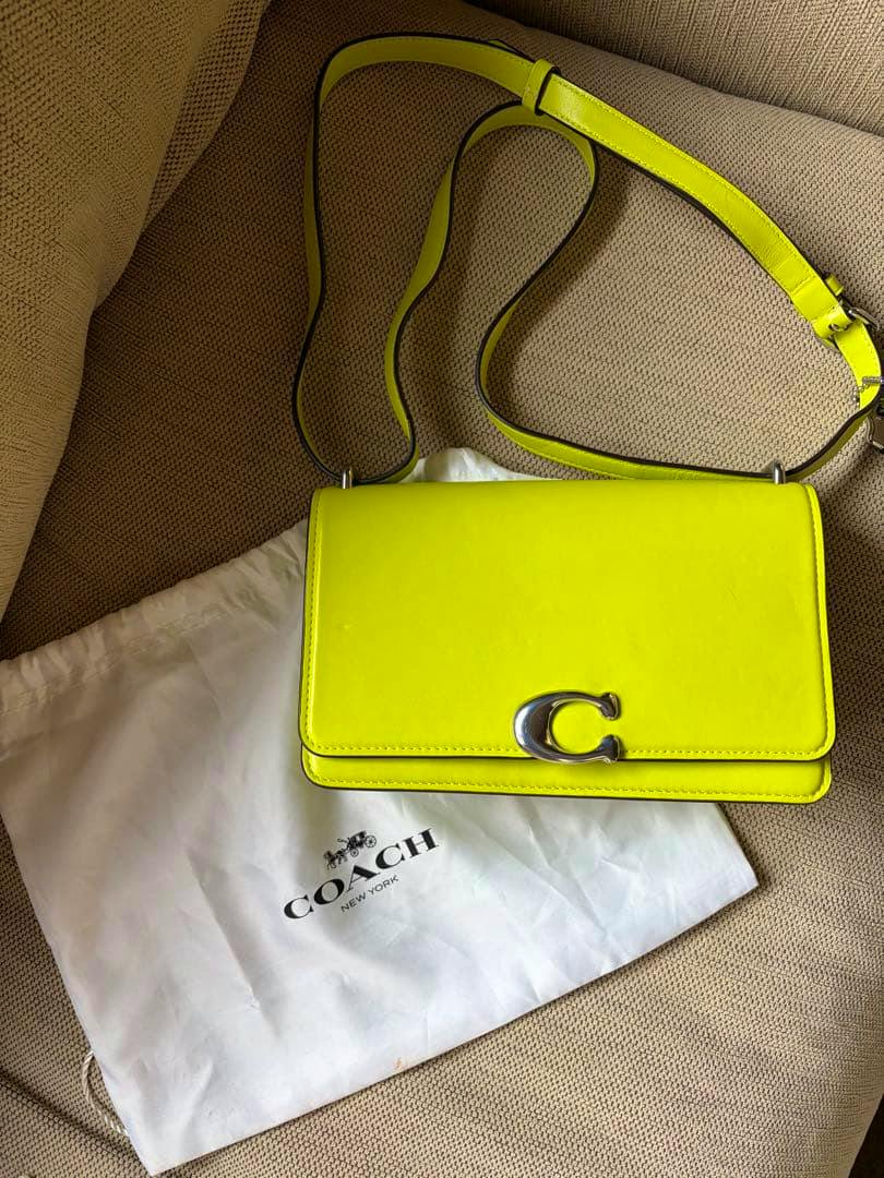 Neon yellow COACH Bandit leather shoulder bag with silver 'C' clasp and adjustable strap on a beige couch above a white Coach dust bag.