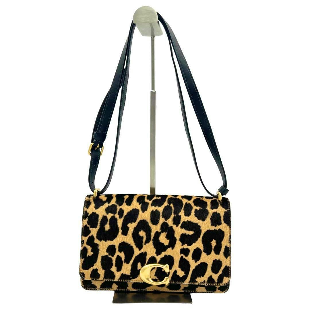 Front view of a COACH Bandit shoulder bag in leopard-print cow leather with black adjustable strap and gold-tone clasp, displayed on a stand.