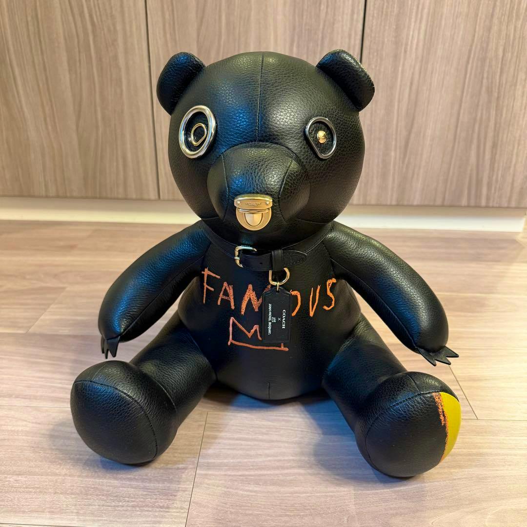 Front view of the Coach Basquiat plush bear showing a black bear with "FAMOUS" painted on its chest and a Coach hang tag.