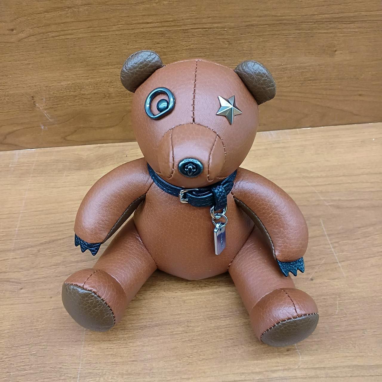 Brown leather COACH bear paperweight with metal star eye and tag sitting on a wooden surface.