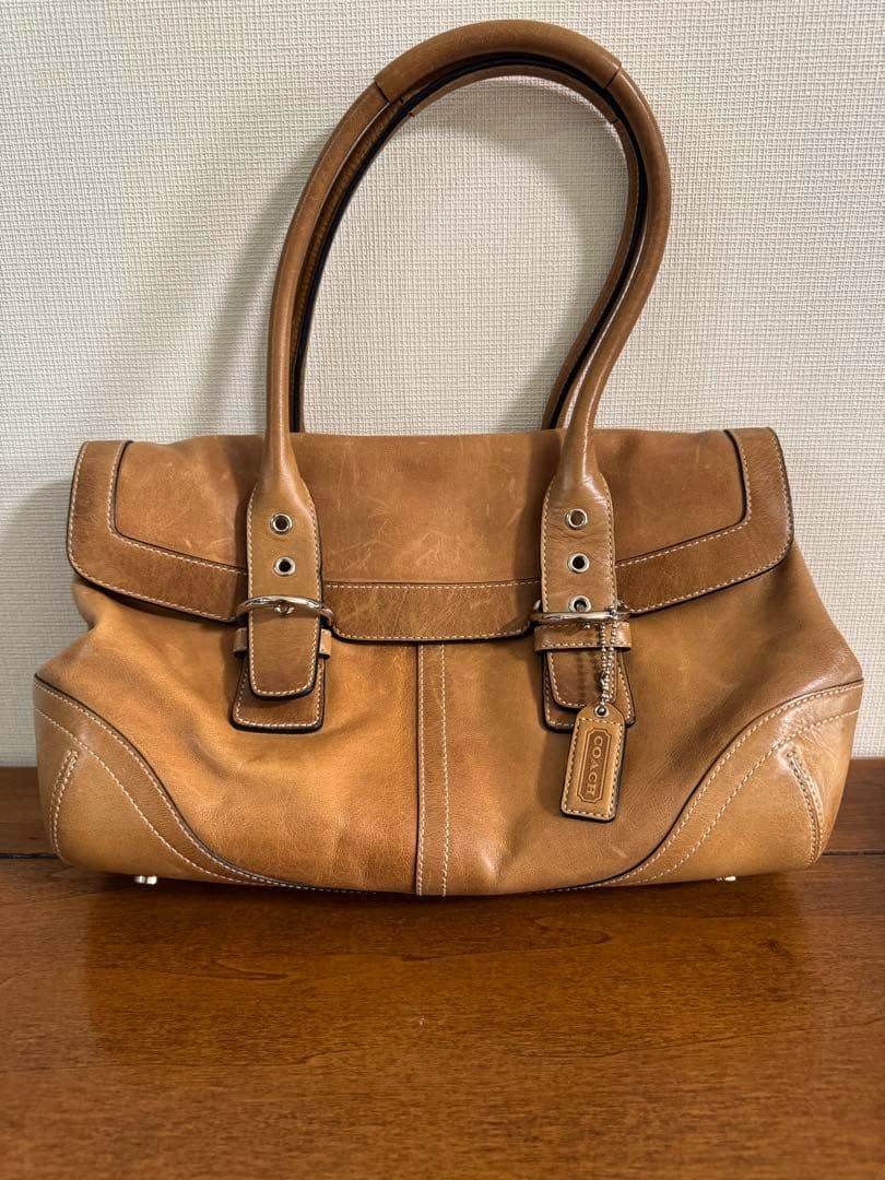 Beige COACH leather flap satchel bag with double handles and front buckle detail on a wooden surface against a textured wall.