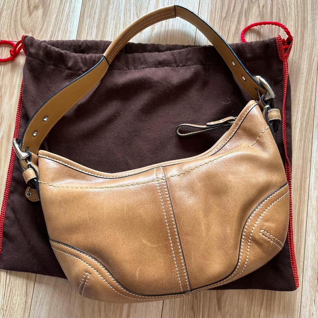 Beige leather COACH shoulder bag laid on a brown dust bag on a wooden floor.
