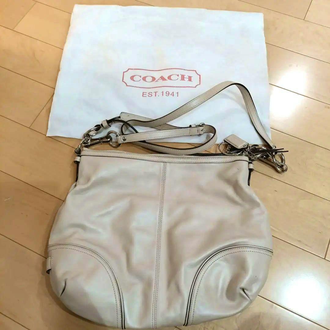 Beige COACH shoulder bag displayed flat with its strap and a Coach dust bag on a wooden floor.