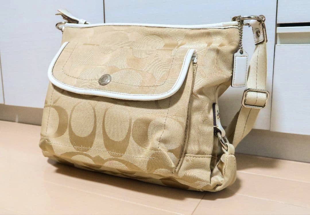 Beige COACH logo-printed canvas shoulder bag shown at a front-right angle with a flap pocket, white trim, and adjustable strap.