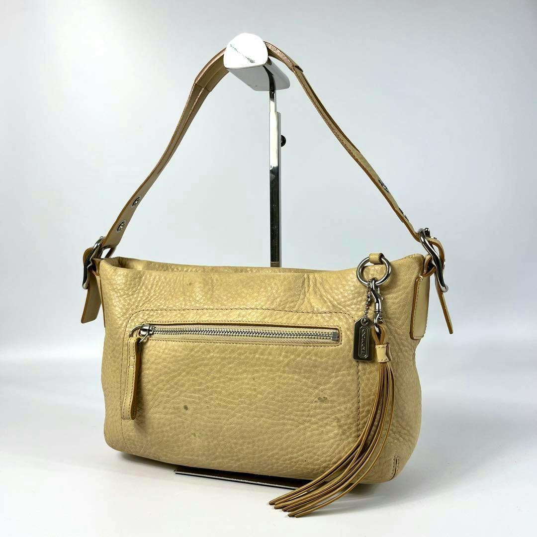 Beige Coach one-shoulder leather bag with front zip pocket and tassel charm on a display stand.