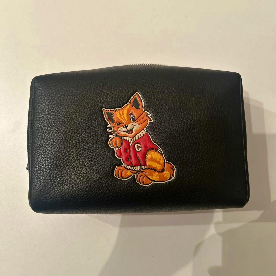 Black cow-leather COACH pouch photographed flat showing an orange cat motif patch on the front.