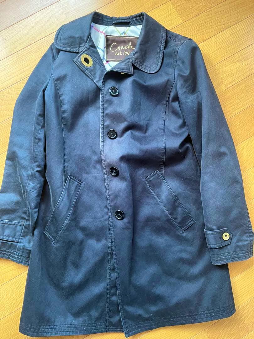 Black Coach women's cotton trench coat laid flat on a wood floor showing the front buttons and pockets.