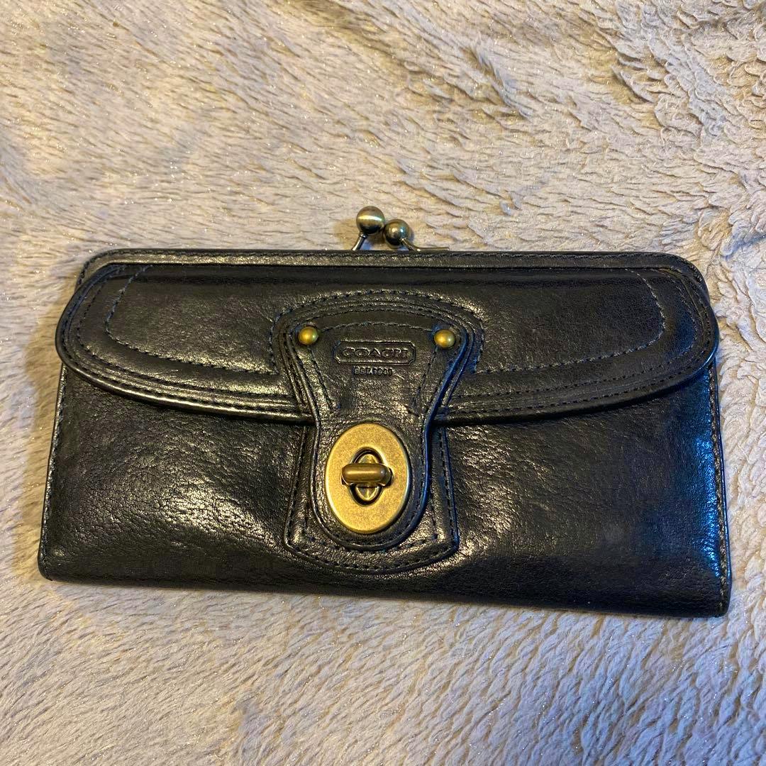Front view of a black Coach long leather wallet with a brass turn-lock closure lying on a textured fabric surface.