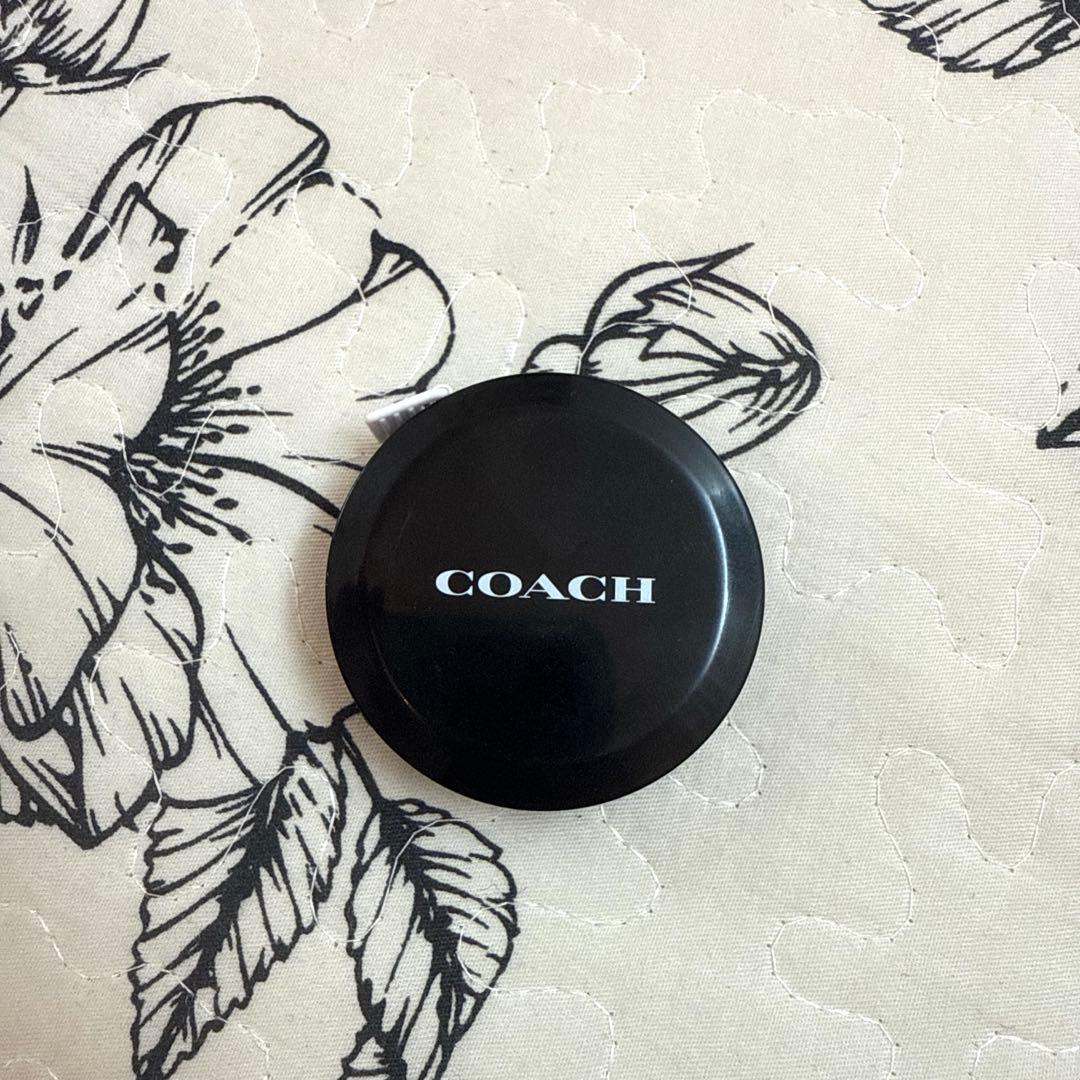 Black round COACH-branded retractable measuring tape case on patterned fabric.