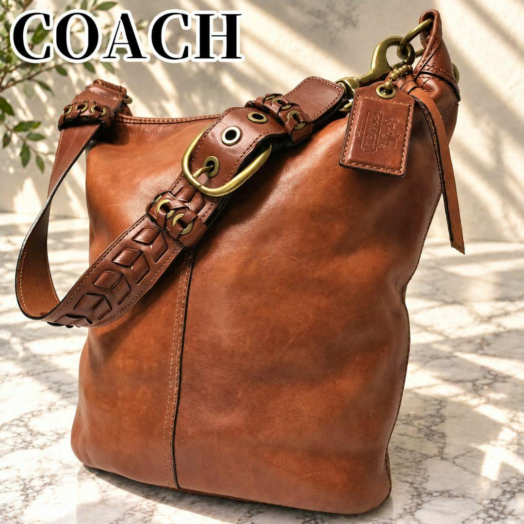 Brown COACH Bleecker duffle shoulder bag in cow leather with braided strap, brass hardware, and leather tag on a marble surface.
